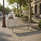 A modern three-seater metal bench with a wire grid back and seat, situated in a sunlit urban plaza with trees, stone paving, and outdoor seating.