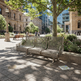 A modern three-seater metal park bench with a woven wire back and seat, situated in a sunny urban plaza with stone paving, trees, and historic buildings in the background.