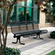 A black metal outdoor bench with a slatted back and seat, placed on a paved walkway in front of a building with large windows and trees in the foreground.