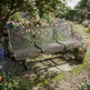 A three-seat garden bench with a woven metal wire frame sits on a stone path in a lush, flower-filled garden under a tree.