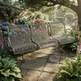 A three-seater metal garden bench with a wire mesh back and seat, shown on a stone path in a lush, sunlit garden with flowers and greenery.