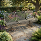 A three-seat metal garden bench with a slatted wire design and curved armrests, placed on a stone path in a lush, sunlit garden with flowers and greenery.