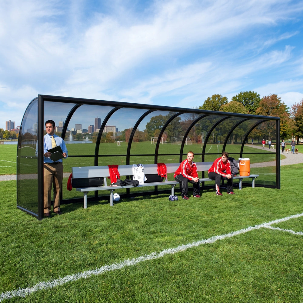 Soccer players and coach in a dugout on a soccer field with clear weather.