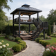 A two-story black metal gazebo with an attached staircase and outdoor seating, set in a landscaped garden with a gravel path leading up to it.