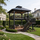 A two-story black wooden gazebo with an attached staircase, situated in a beautifully landscaped garden with a stone pathway, manicured hedges, and large white villas in the background.