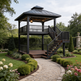A two-tiered black wooden gazebo with an attached staircase, situated in a beautifully landscaped garden with a gravel path leading up to it.