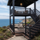 A coastal two-story screened porch with dark stained wood framing and staircase, furnished with a sofa and coffee table, overlooking the ocean and rocky cliffs.