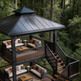A large multi-level outdoor pavilion with a dark gray metal roof and black railings, furnished with outdoor seating and a staircase leading down into wooded terrain.