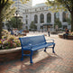 A bright blue slatted metal park bench in a city plaza, with brick paving, flower beds, trees, and a large classical building in the background. People are seated and walking nearby, so this is a Lifestyle image rather than a clean Product/Packaging shot.