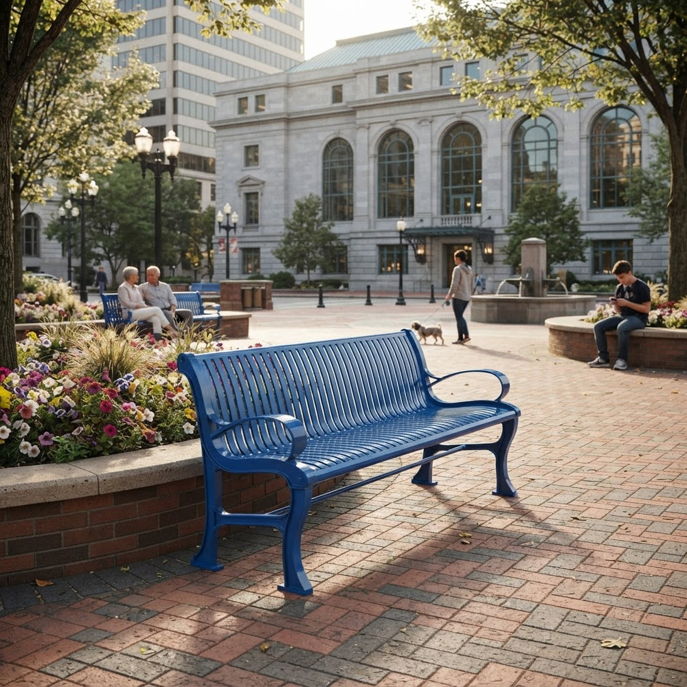A bright blue slatted metal park bench in a city plaza, with brick paving, flower beds, trees, and a large classical building in the background. People are seated and walking nearby, so this is a Lifestyle image rather than a clean Product/Packaging shot.