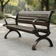 A dark brown wooden park bench with ornate metal legs and armrests, shown on a paved path in a park setting.
