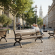A wooden park bench with dark metal arms and legs sits on a cobblestone plaza in a city square, with an elderly couple seated on it. Trees, historic buildings, and a street lamp are visible in the background.