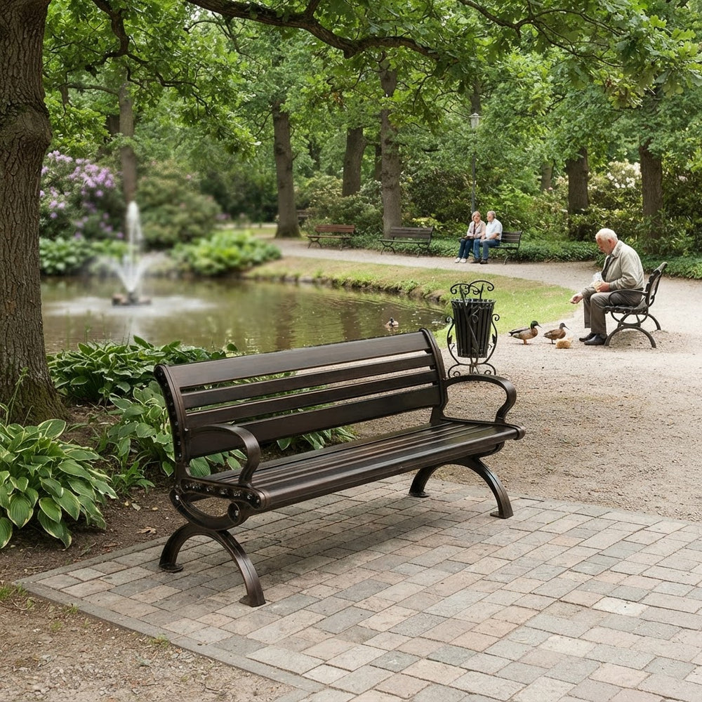 A dark brown wrought-iron park bench with wooden slats is shown in a lush green park setting next to a pond with a fountain, with people and ducks in the background.