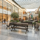 A modern outdoor shopping plaza with dark metal benches, a glass roof, and visible storefronts including Céline and Hugo. A colorful abstract sculpture stands near the center, with shoppers walking in the background.