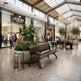 A modern shopping mall interior with wooden park benches, large potted green plants, a stainless steel trash can, and shoppers walking or sitting in a bright, upscale atrium. Visible storefront signs include Aurora Jewelry and Cafe Terrace.