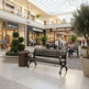 A dark brown wooden park bench with curved arms and legs stands in a modern shopping mall atrium with a glass roof, surrounded by potted trees, planters, and other shoppers.