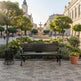 A dark brown wooden park bench with curved black metal arms sits on a paved plaza in a beautifully landscaped town square with a fountain, trees, and historic buildings in the background.