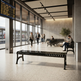 A modern airport terminal interior with long black slatted benches, travelers walking and sitting near large windows, a departure board on the left, and a clean architectural background.