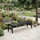 A black slatted outdoor bench with curved legs, placed on a light stone patio beside a landscaped garden bed with flowers, greenery, and a garden hose in a park-like setting.