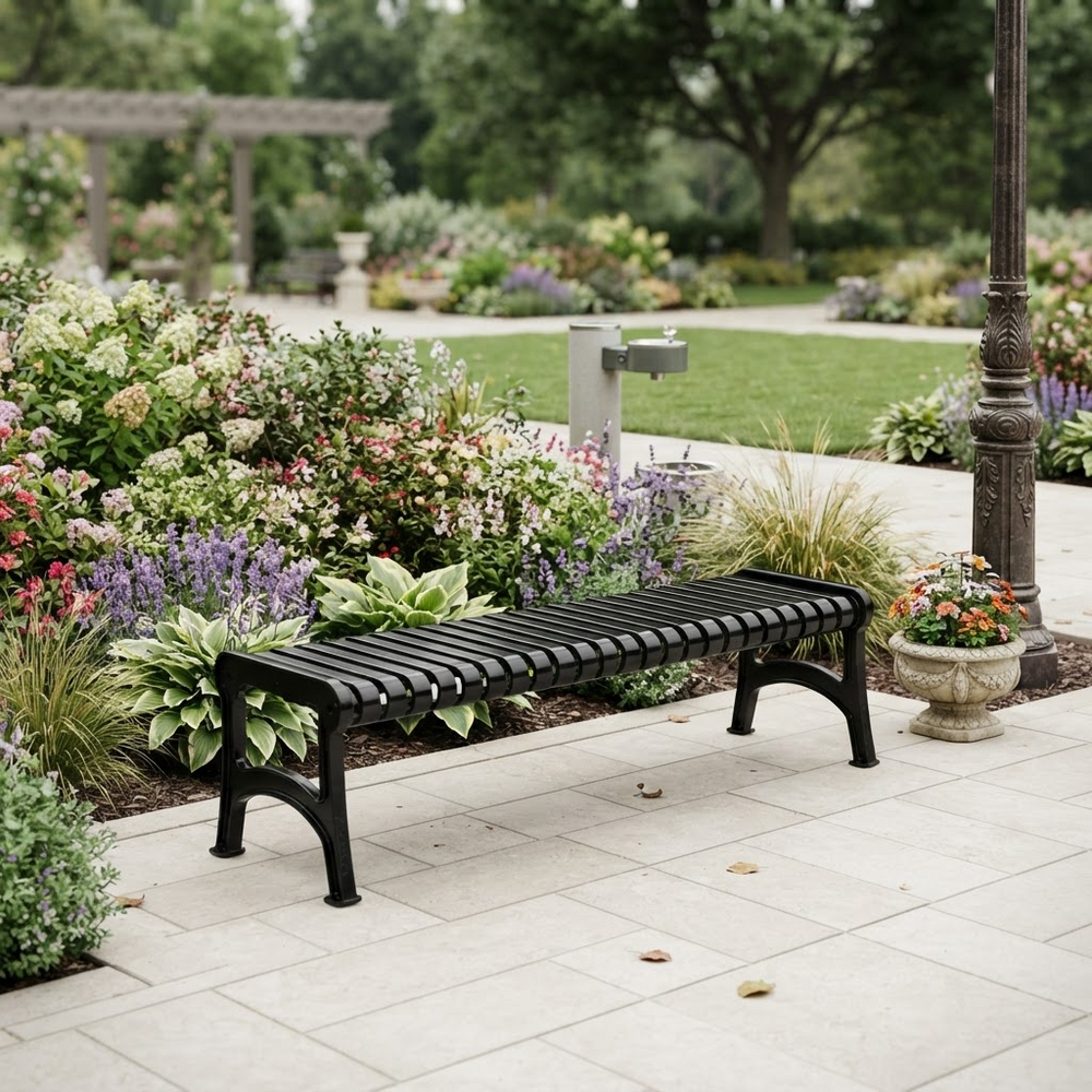 A black slatted outdoor bench with curved legs, placed on a light stone patio beside a landscaped garden bed with flowers, greenery, and a garden hose in a park-like setting.