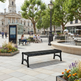 A black metal slat bench in a public square, with people sitting nearby, trees, a fountain, and a city building in the background.