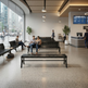 A modern, spacious airport or transit center waiting area with people sitting on black slatted benches near large floor-to-ceiling windows. The space has a polished terrazzo floor, hanging pendant lights, potted plants, a reception desk, and a departure board on the wall.