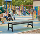A black slatted park bench in the foreground at a children's playground, with kids, a stroller, colorful play structures, and a playground sign in the background.