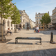 A wide urban plaza with stone paving, classical buildings, trees, benches, and several pedestrians under a blue sky.