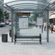 A modern glass and metal bus stop shelter with a black slatted bench, a trash can, and route information boards, set on a paved city sidewalk with blurred pedestrians and street activity in the background.