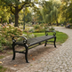A black metal park bench with curved armrests sits on a cobblestone path in a lush green park with trees, flowers, and distant fountain visible.