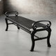 A black wrought-iron outdoor bench with curved scrollwork ends and a slatted seat, photographed on a concrete floor against a softly blurred brick wall.