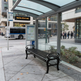 A modern glass and metal bus stop shelter with a black slatted bench on a city sidewalk. A City Transit bus is visible in the background, along with buildings, street elements, and a few pedestrians.