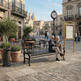 An elderly couple sits on a black metal bench in a sunny, historic European-style plaza, reading a newspaper. The scene features stone buildings, potted olive trees, outdoor cafe tables, a vintage street clock, and a gold-framed information board in the background.