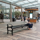 A black metal bench sits in a transit station waiting area, with two people seated on it. A city transit hub kiosk, glass-covered roof, statue, and walking passengers are visible in the background.