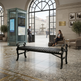 A black metal slatted bench sits in a grand transit station lobby with large arched windows, a glass transit kiosk, seated passengers, and a polished marble floor.