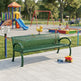 A green slatted metal park bench in a paved playground area, with a colorful children's playground, swings, and trees in the background. A small toy truck and a blue bottle are on the pavement in front of the bench.