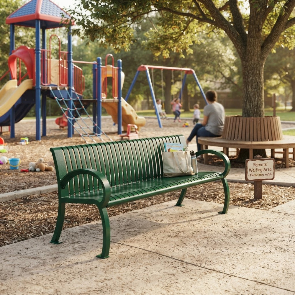A green metal park bench with curved armrests sits on a concrete path in a playground park. A white tote bag rests on the bench, and a child is sitting on a wooden bench in the background near a playground and trees.
