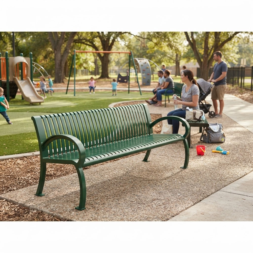 A green metal park bench sits on a paved walkway in a sunny playground setting, with children playing in the background and a woman sitting nearby on a small table.