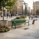 A green slatted public bench in a modern urban plaza, surrounded by flower beds, a tree in a large metal planter, and pedestrians walking in the background between historic and contemporary buildings.