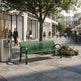 A green slatted outdoor bench on a paved shopping plaza sidewalk, with a tree, storefronts, pedestrians, a flower bed, and a recycling bin in the scene.