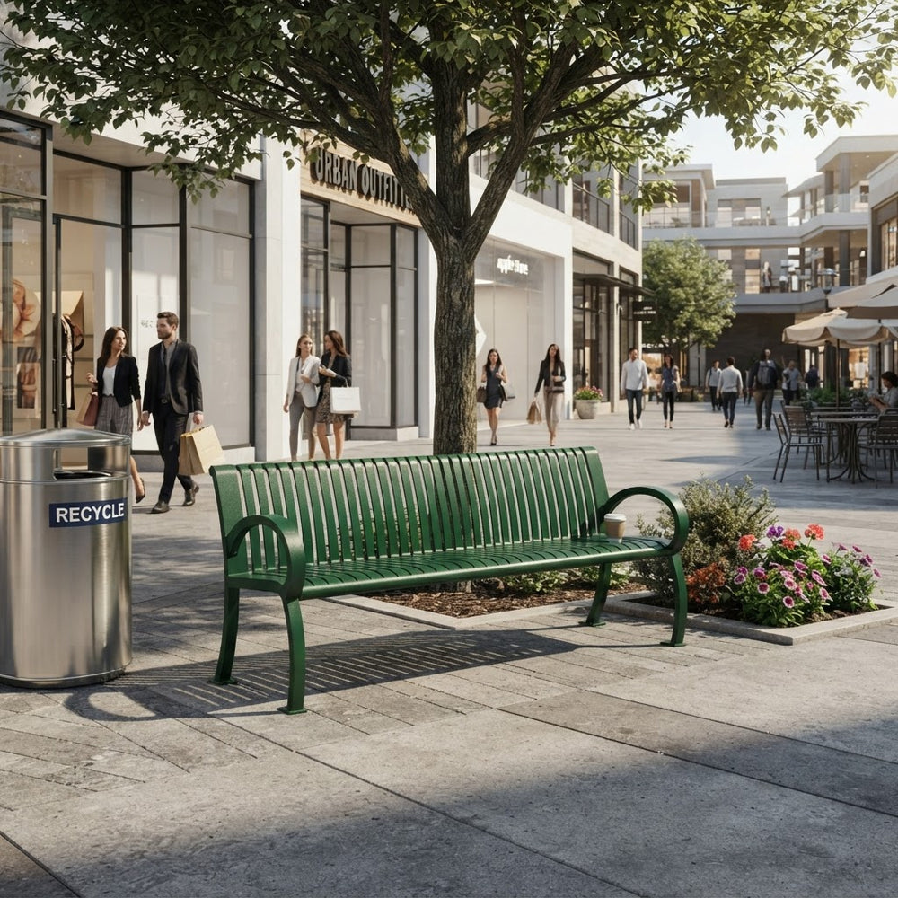 A green slatted outdoor bench on a paved shopping plaza sidewalk, with a tree, storefronts, pedestrians, a flower bed, and a recycling bin in the scene.