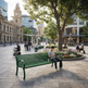 A green outdoor park bench in a city plaza, with a young man sitting on it and reading a book. The scene includes other people, trees, flowers, and urban buildings in the background.