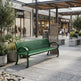 A green metal slat bench in an outdoor shopping plaza, with storefronts, potted plants, and a few pedestrians visible in the background.