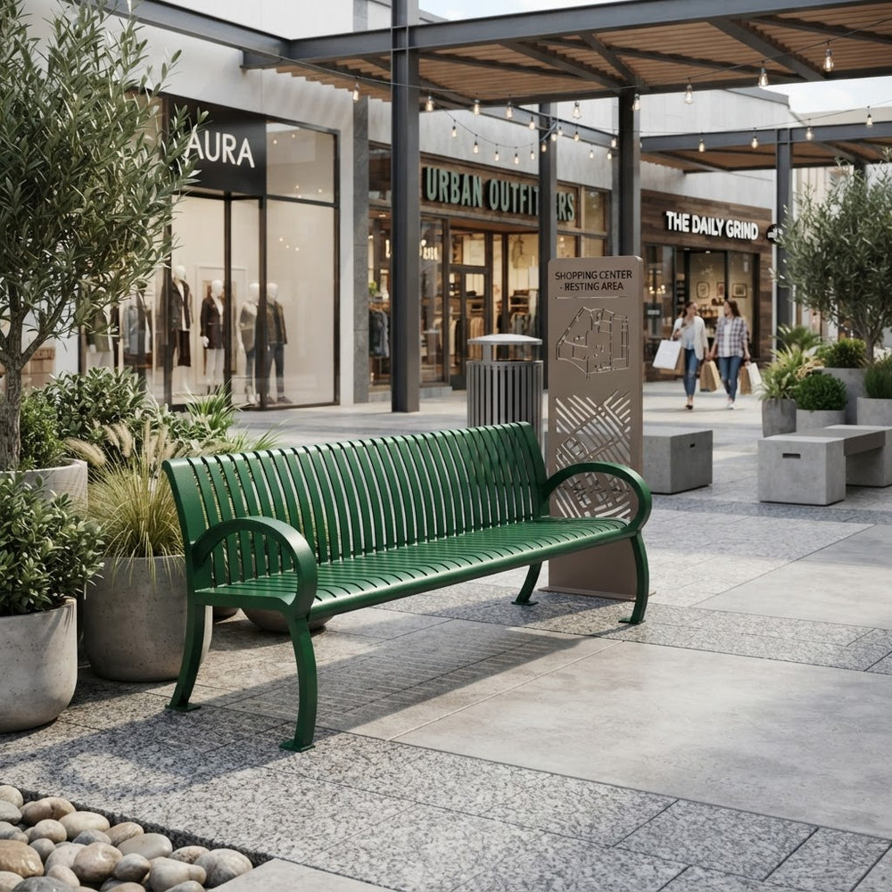 A green metal slat bench in an outdoor shopping plaza, with storefronts, potted plants, and a few pedestrians visible in the background.