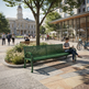 A green metal park bench in a sunny urban plaza, with a woman sitting on one end reading a book. Pedestrians walk around the plaza, with a modern glass-front building and a historic clock tower visible in the background.