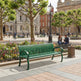 A green metal park bench in a city plaza, with people sitting nearby, trees, historic buildings, and a black wrought-iron fence in the background.