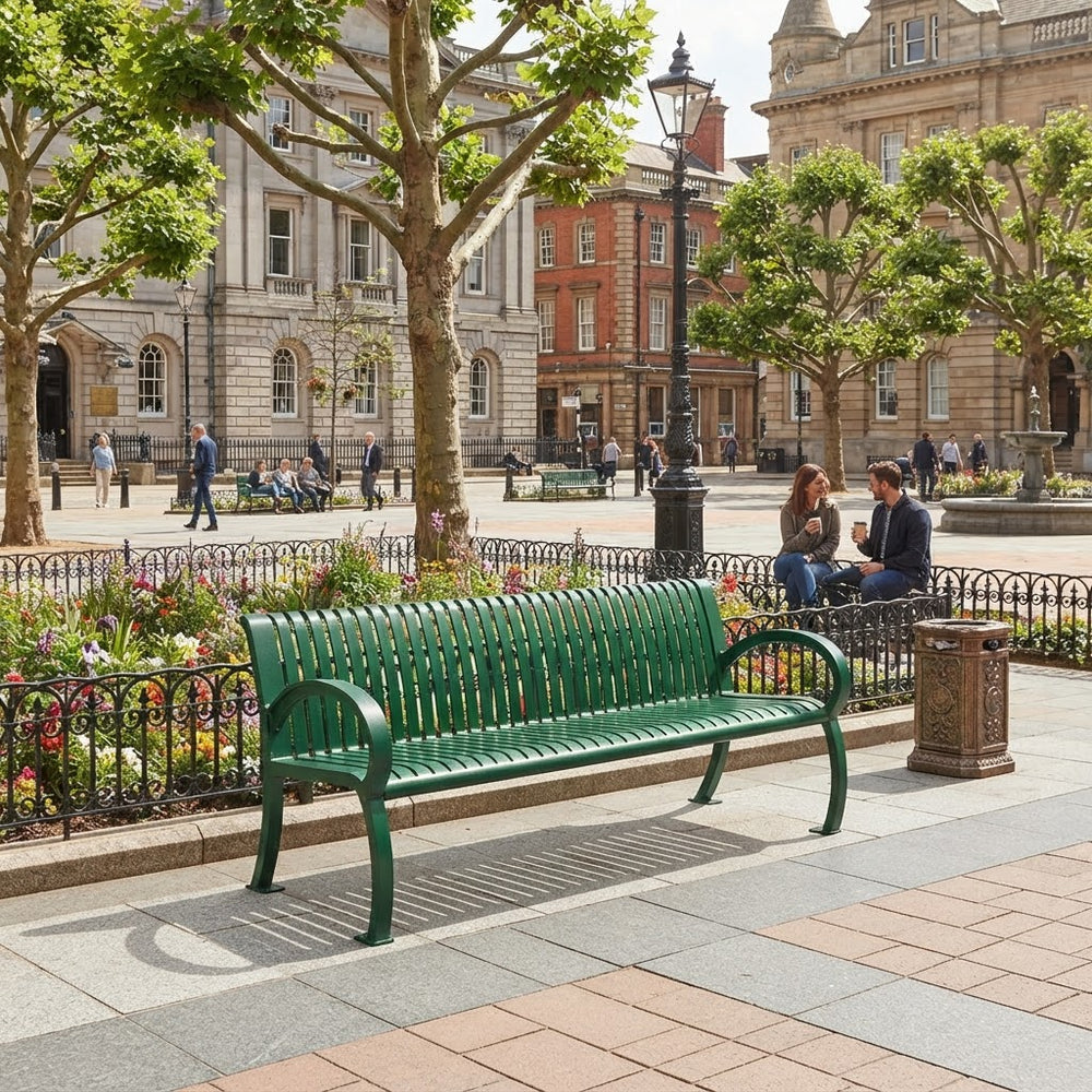 A green metal park bench in a city plaza, with people sitting nearby, trees, historic buildings, and a black wrought-iron fence in the background.