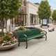 A green metal park bench with curved armrests sits on a paved outdoor plaza in front of storefronts. Two people are seated on the bench, and large flower planters, trees, trash cans, and a digital map kiosk are visible in the surrounding shopping-area setting.