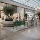 A modern green slatted bench sits on a concrete base in a spacious, sunlit indoor shopping mall. Two people are seated on the bench, and the background features a boutique with large glass windows, potted olive trees, and a glass-roofed ceiling.