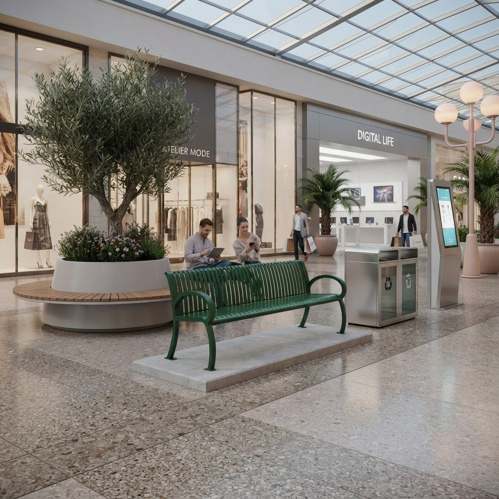 A modern green slatted bench sits on a concrete base in a spacious, sunlit indoor shopping mall. Two people are seated on the bench, and the background features a boutique with large glass windows, potted olive trees, and a glass-roofed ceiling.